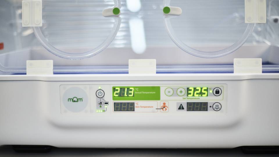 A close up of a neonatal incubator, showing two portholes and a control panel bearing the MoM incubator logo